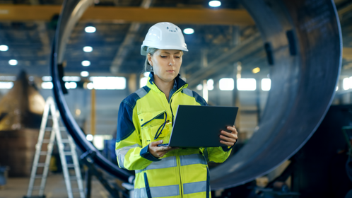 A woman in a hard hat and safety vest holding a laptop, ready to work on a construction site.